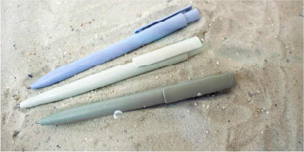 Drei Kugelschreiber aus recycelten PET-Flaschen liegen am Strand im Sand.