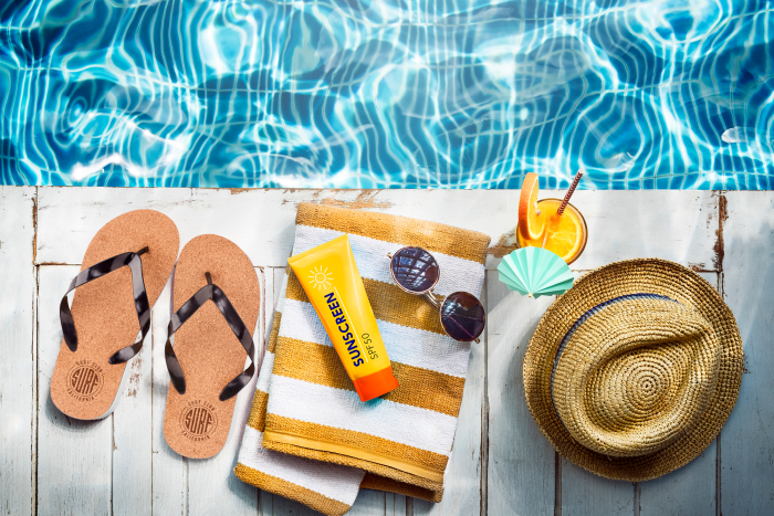 Verschiedene Werbeartikel für den Sommer liegen an einem Pool. Zehensandalen, Sonnencreme, Sonnenbrille und ein Strohhut sind perfekte Geschenke für die Sommerzeit.